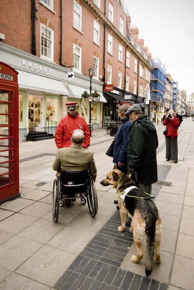 Eric Sayce with Wills is standing with his back to the camera. There is a man in a wheel chair to his left and they are talking to a man in a red cap and coat who is a West End guide; this is a newly established company to help people to find their way around the area.